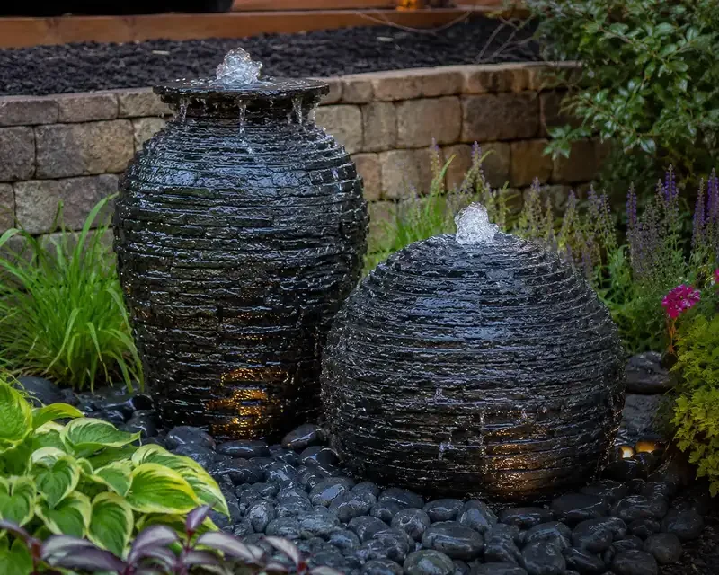 Two black stone water fountains in a garden surrounded by green plants and purple flowers, with a brick wall in the background.
