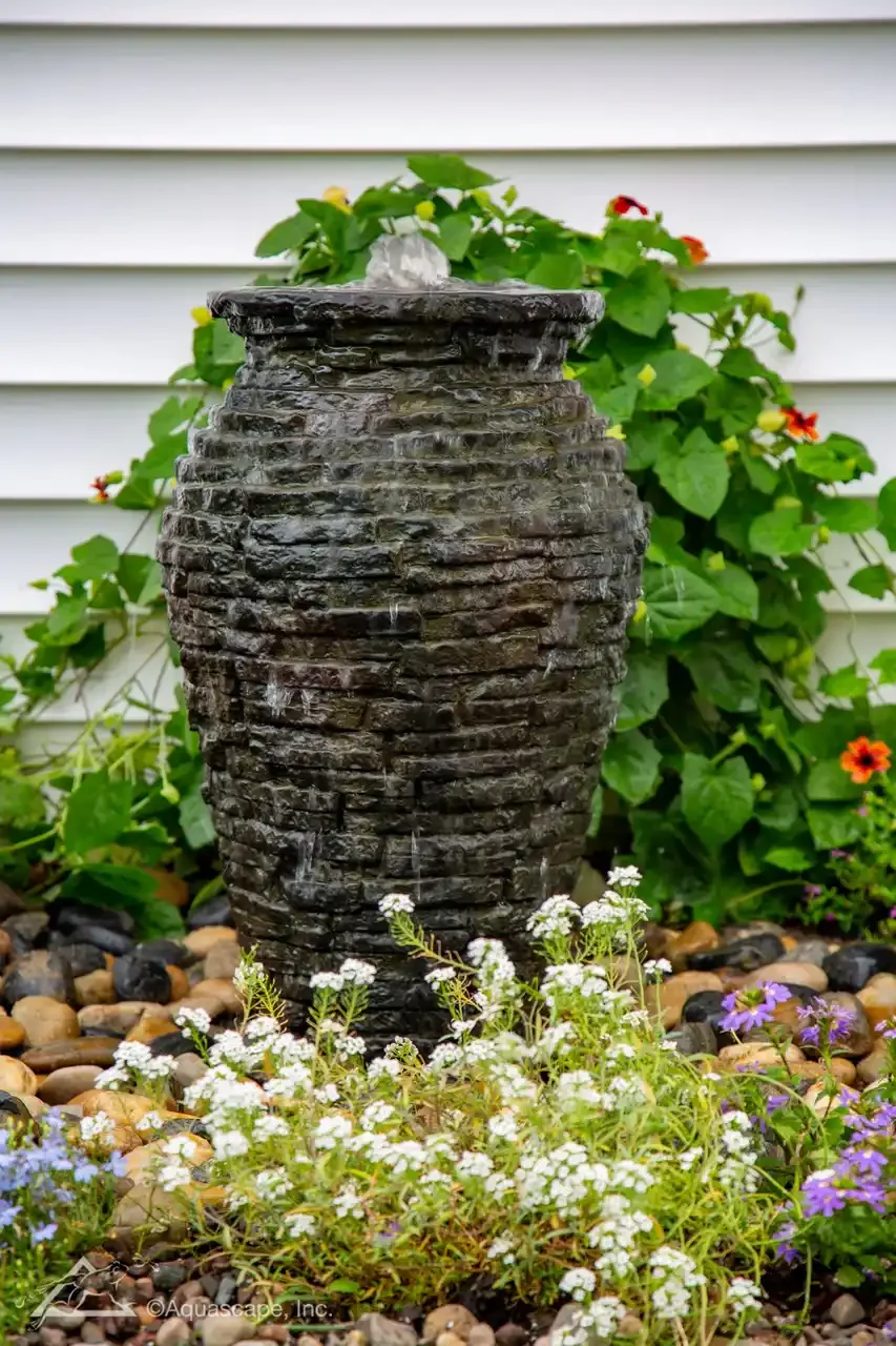A black textured stone outdoor fountain with water flowing from the top, surrounded by colorful flowers and green foliage, with a white house siding in the background.