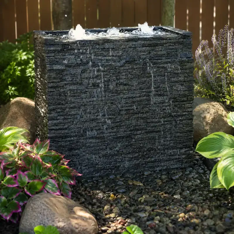 A black square water fountain with water flowing from the top, surrounded by green plants and rocks.