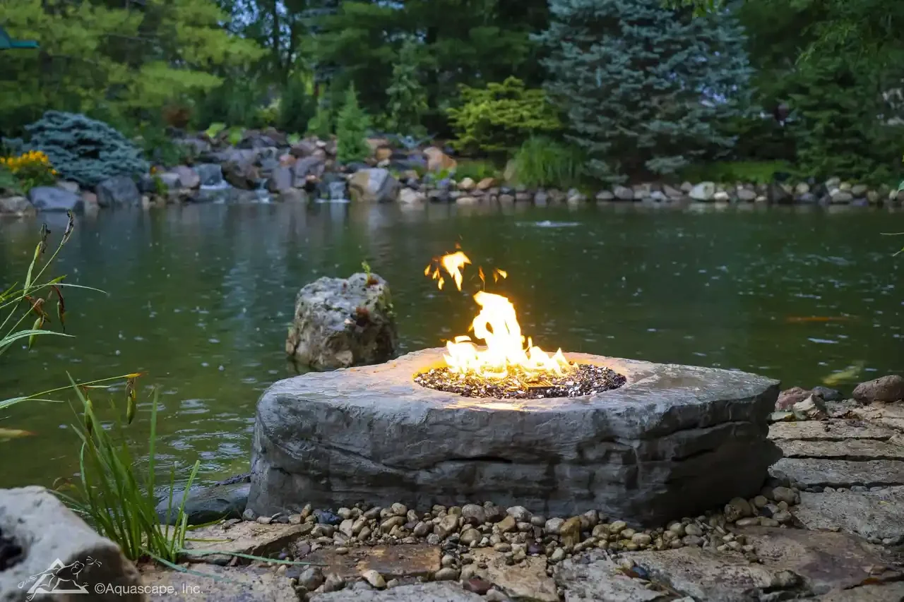 A fire pit with flames by a pond surrounded by rocks and lush green trees.