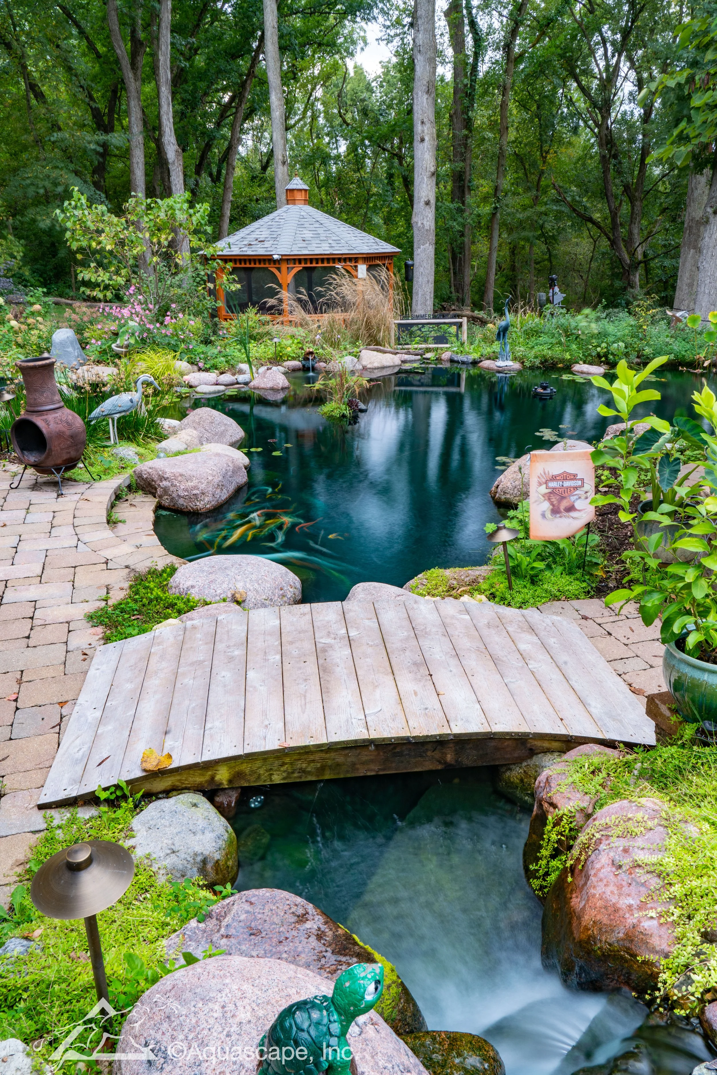 Aquascape ecosystem pond with a decorative wood bridge, natural stone edging, and lush backyard landscaping.