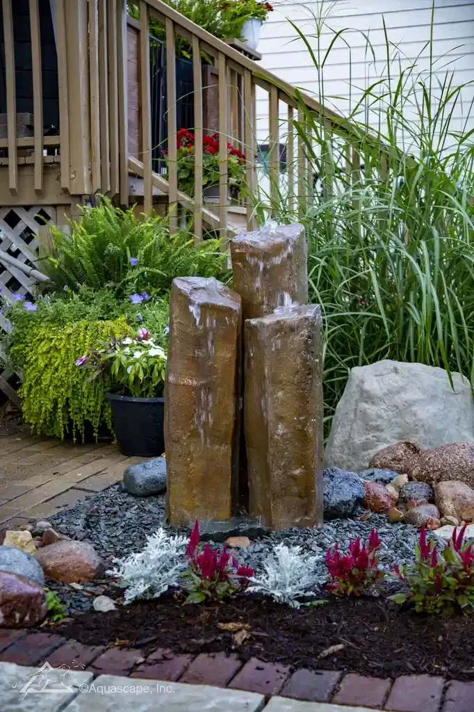Water feature with four stacked brown stone pillars, water cascading down their sides, surrounded by red, white, and green plants, with a wooden deck and house siding in the background.