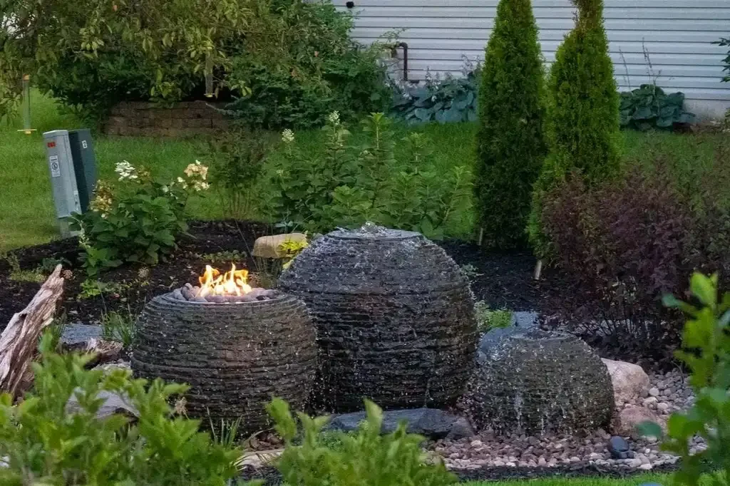 Garden with three large black textured water features, a small fire in one of the features, surrounded by plants, shrubs, and trees, with a house and grass area in the background.