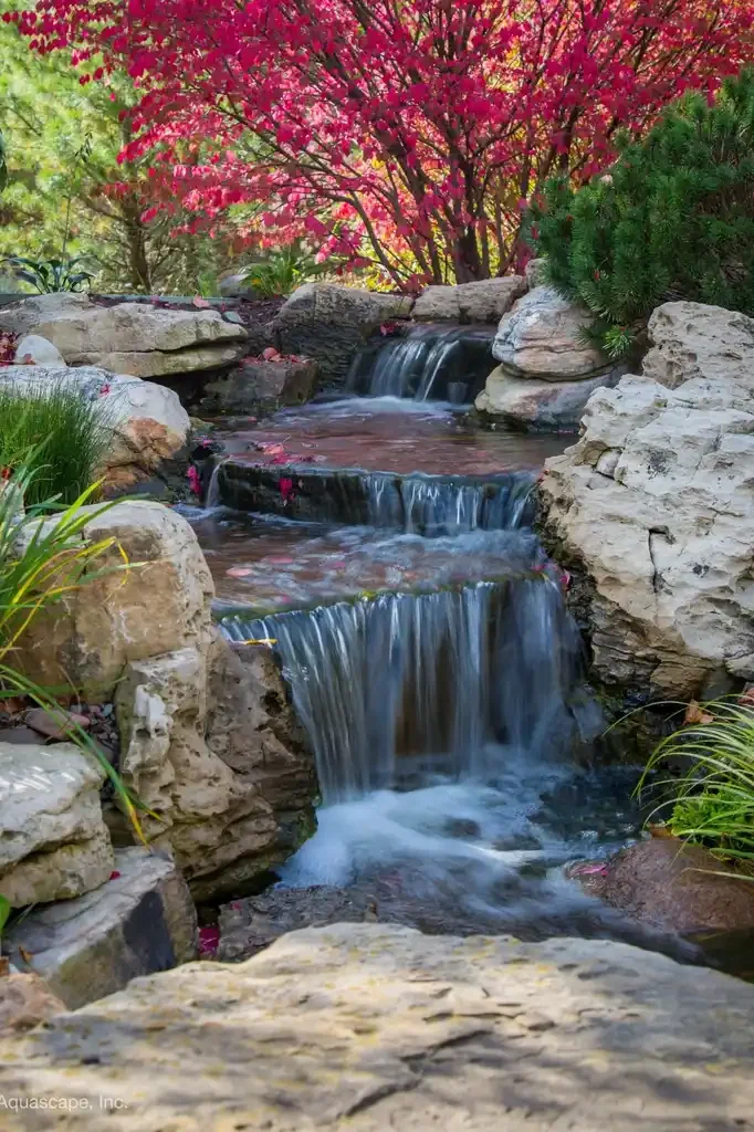 A small cascading waterfall flowing over rocks with a pink flowering tree and green bushes in the background.