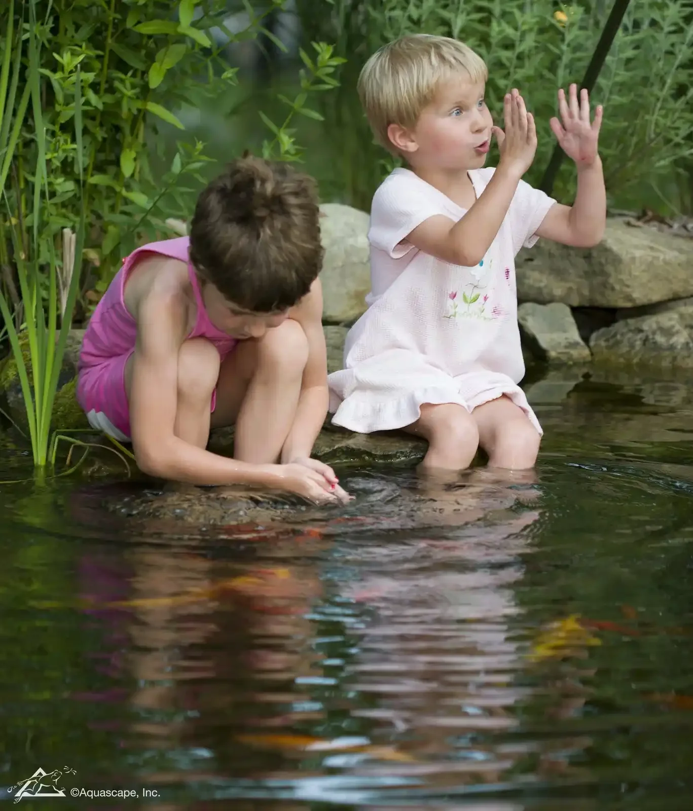 Two young children, a girl and a boy, sitting by the edge of a pond with lush green plants in the background. The girl is squatting and playing with something in the water, while the boy, dressed in a white dress, is sitting with his legs in the water, gesturing with his hands as if talking or explaining something.