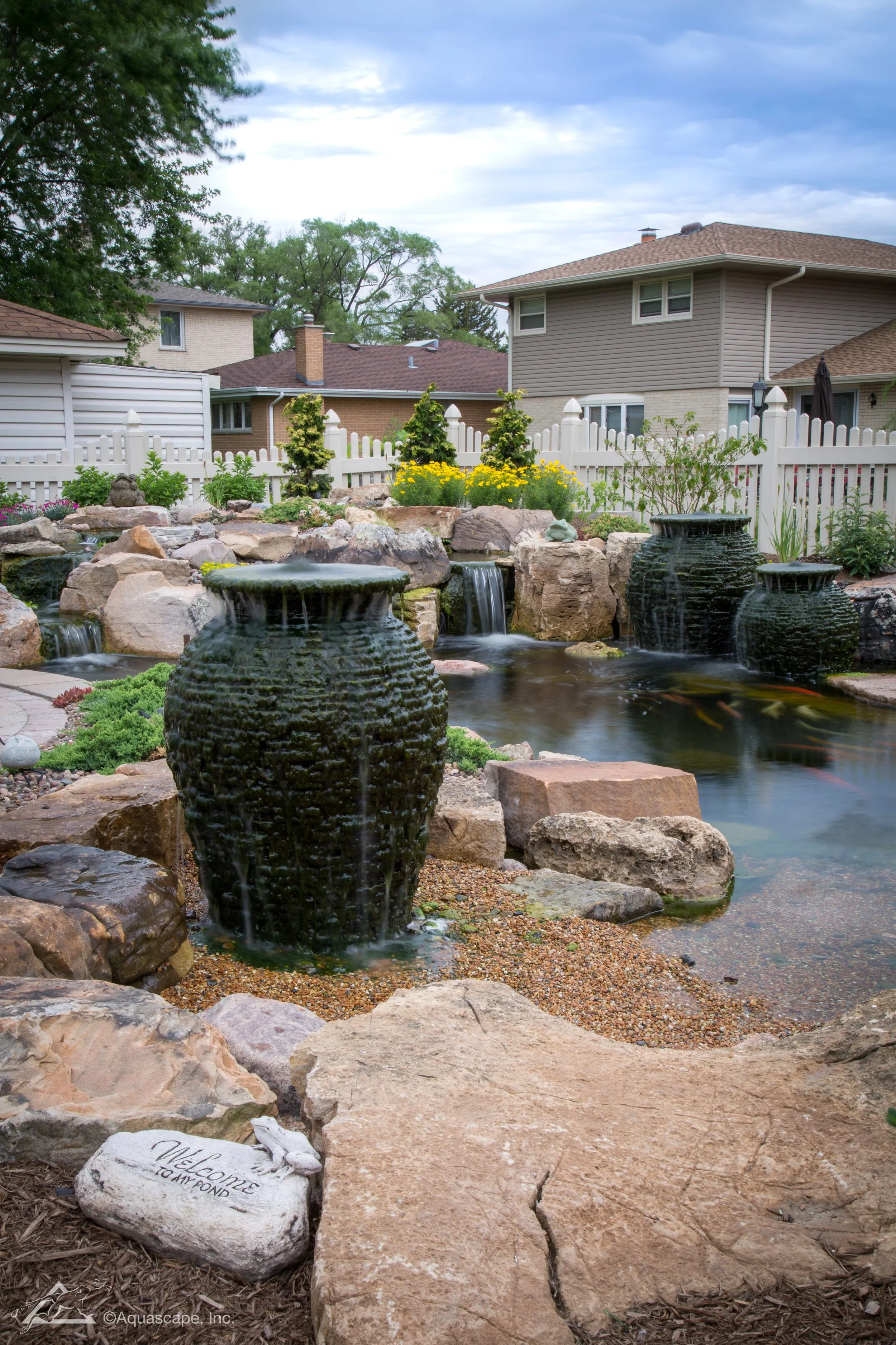Backyard with a pond, rocks, and water feature, surrounded by a white fence and houses, under cloudy sky.