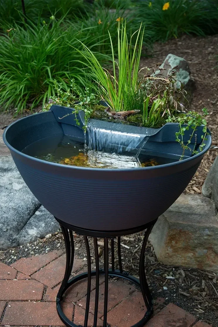 A decorative outdoor water fountain with plants growing in a basin, positioned on a metal stand, surrounded by rocks, bricks, and garden plants.
