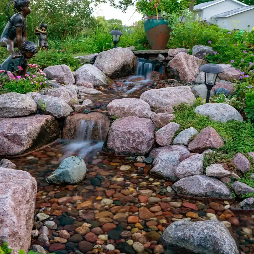 A backyard garden with a small man-made stream flowing over rocks, surrounded by plants and flowers, with two children playing nearby and garden lanterns along the stream.