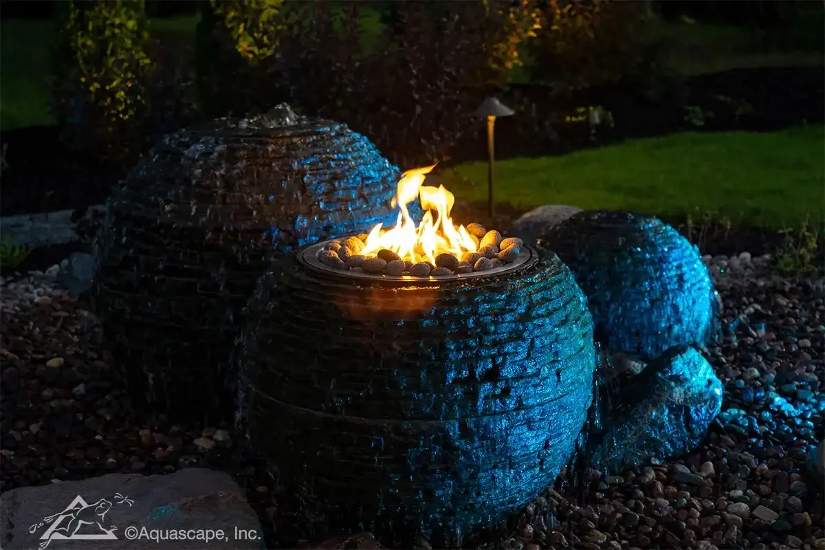 A decorative outdoor water fountain with a fire feature in the center, surrounded by smooth stones, night scene with garden in the background.