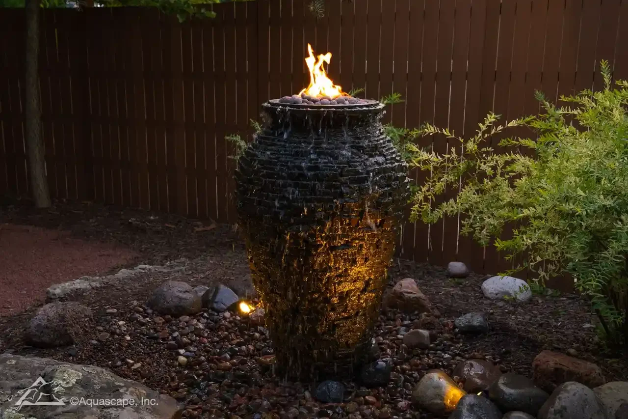A decorative outdoor water fountain with fire on top, surrounded by rocks in a garden with a brown wooden fence and greenery.