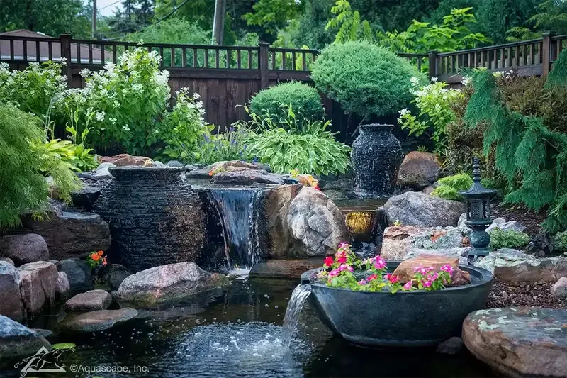 A landscaped backyard pond with cascading water features, surrounded by lush green plants and rocks, including a decorative black lantern and a black basin with pink flowers.