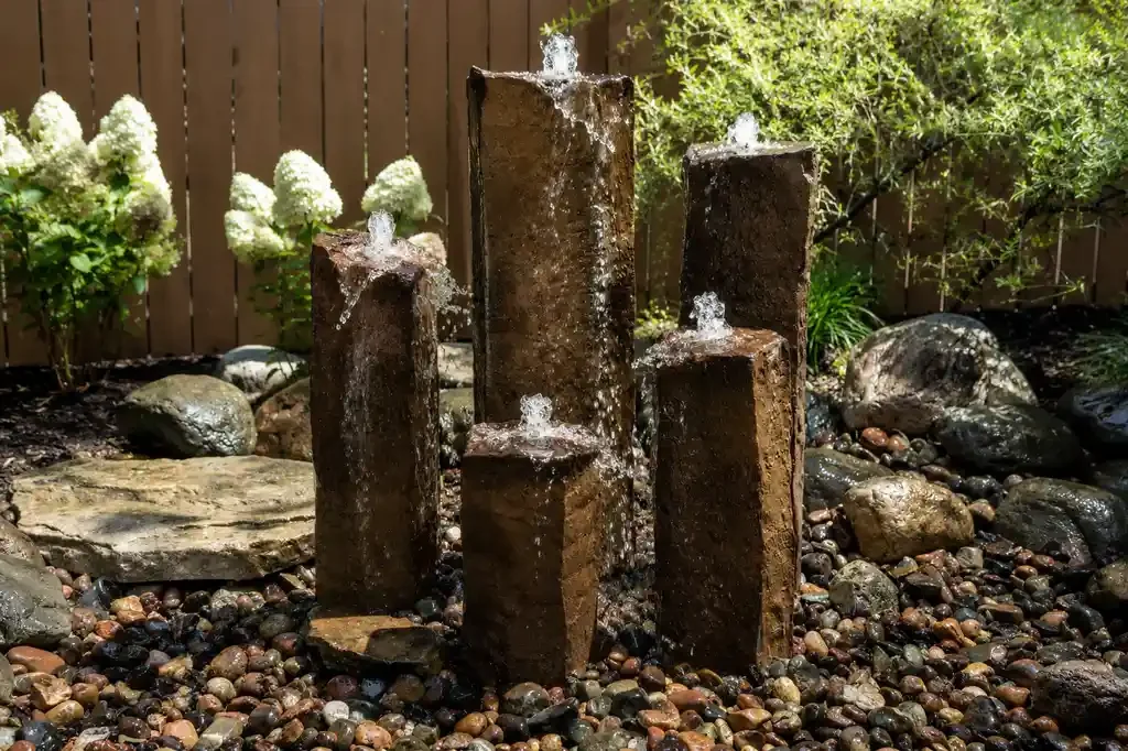 Decorative outdoor water fountain with five rusty metal pillars of varying heights, water flowing from the top of each into a bed of small multicolored pebbles, surrounded by rocks, plants, and flowers.