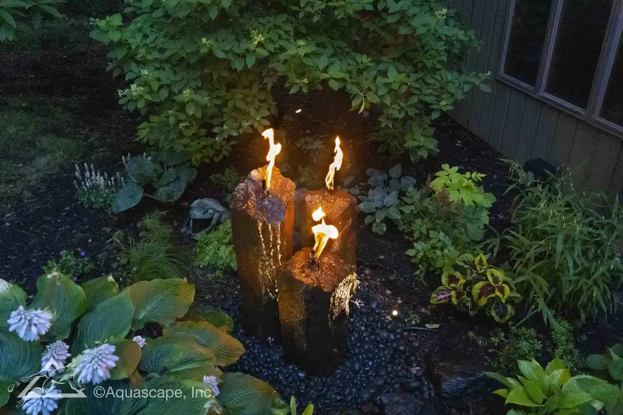 Three decorative stone pillars with flames on top, situated in a landscaped garden with various green plants and bushes.