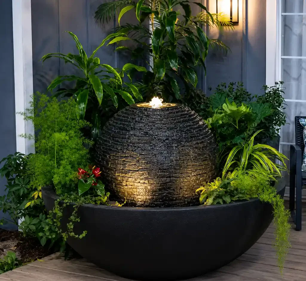 An outdoor arrangement with green plants surrounding a black water fountain with a spherical top that has water flowing down its textured surface, illuminated by soft lighting at night.