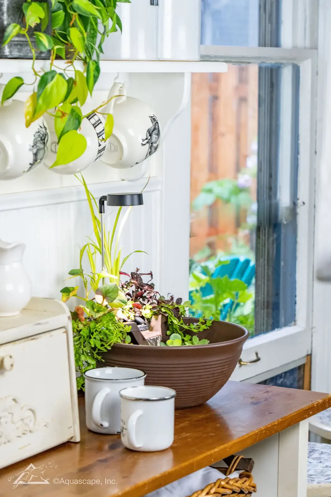 A windowsill and table with potted and hanging plants, two white mugs, a white decorative box, and a view of a garden outside with colorful chairs and greenery.