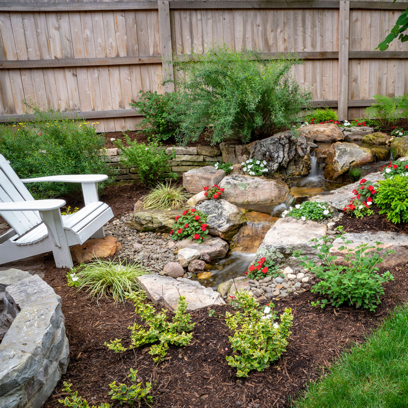 Aquascape-style 6-foot pondless waterfall stream featuring natural stone waterfalls, colorful landscaping, and a relaxing outdoor living space.