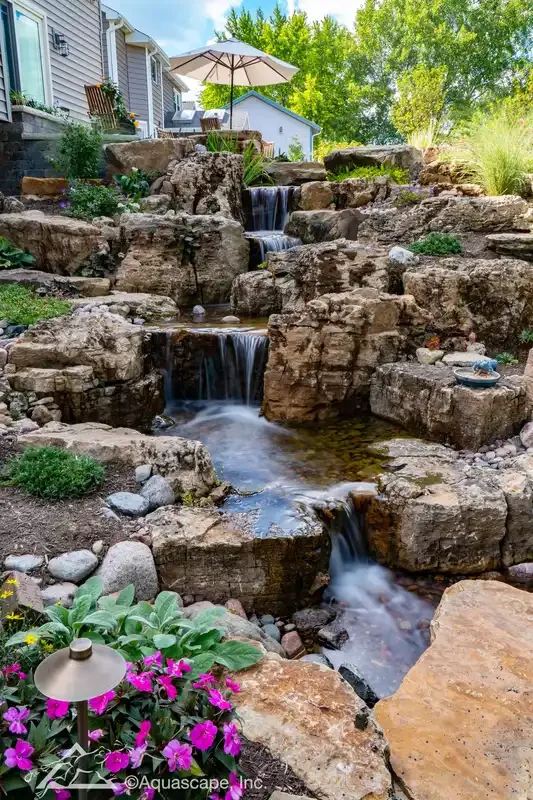 A multi-tiered backyard waterfall with rocks, surrounded by plants and flowers, with a patio umbrella and seating area in the background.