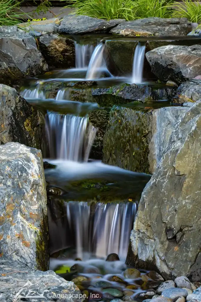 Small waterfall flowing over rocks in a garden setting with green plants in the background.
