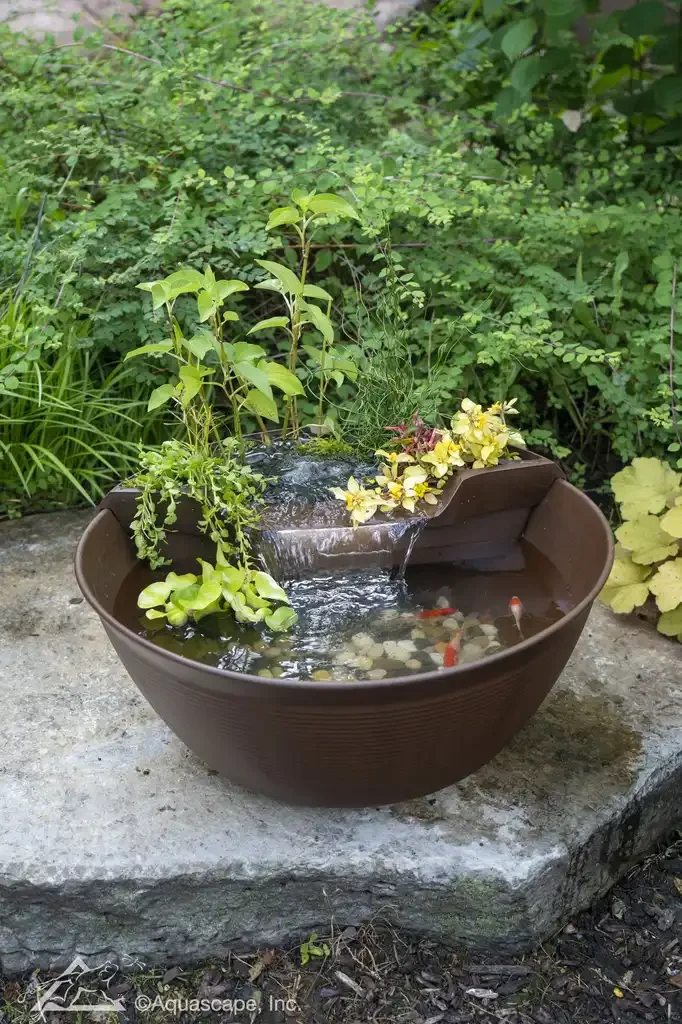 A garden water feature in a brown pot with small cascading waterfall, aquatic plants, and fish, set on a stone surface with green foliage in the background.