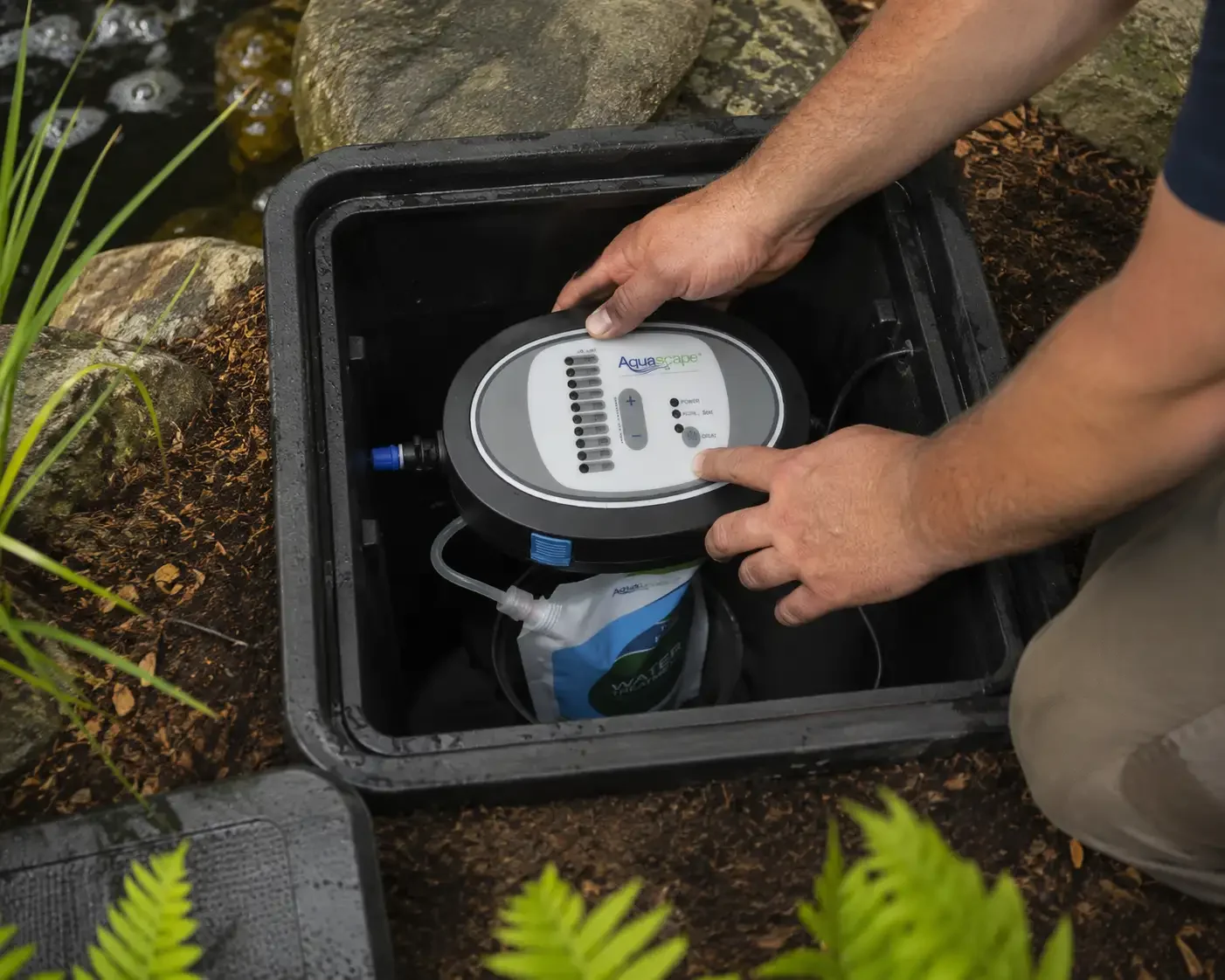 A person installing or servicing an Aqua scape water filtration system in a black container near a pond with rocks and plants.