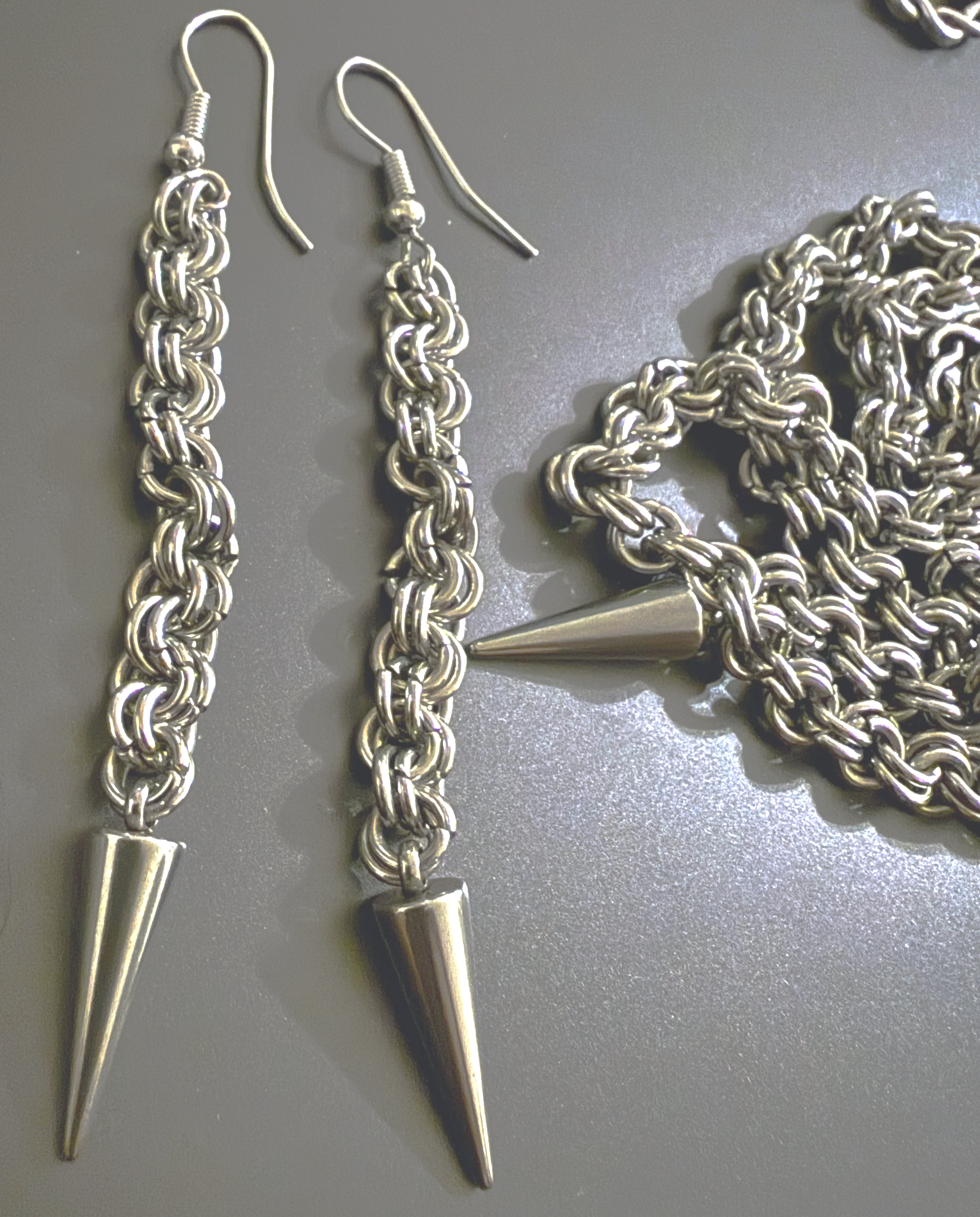 Handmade chainmail earrings with silver spike pendants next to a matching chainmail necklace on a dark background.
