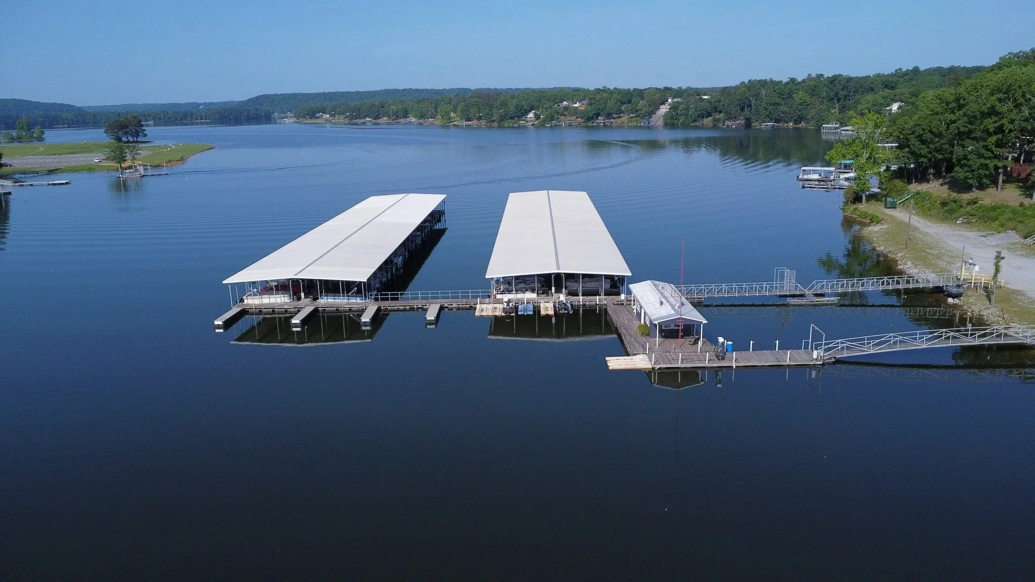 Aerial view of a lakeside marina with two covered boat slips housing over 100+ boats, a dock, and surrounding trees and houses. These boat slips are rented out through Smith Lake Marina and Resort.