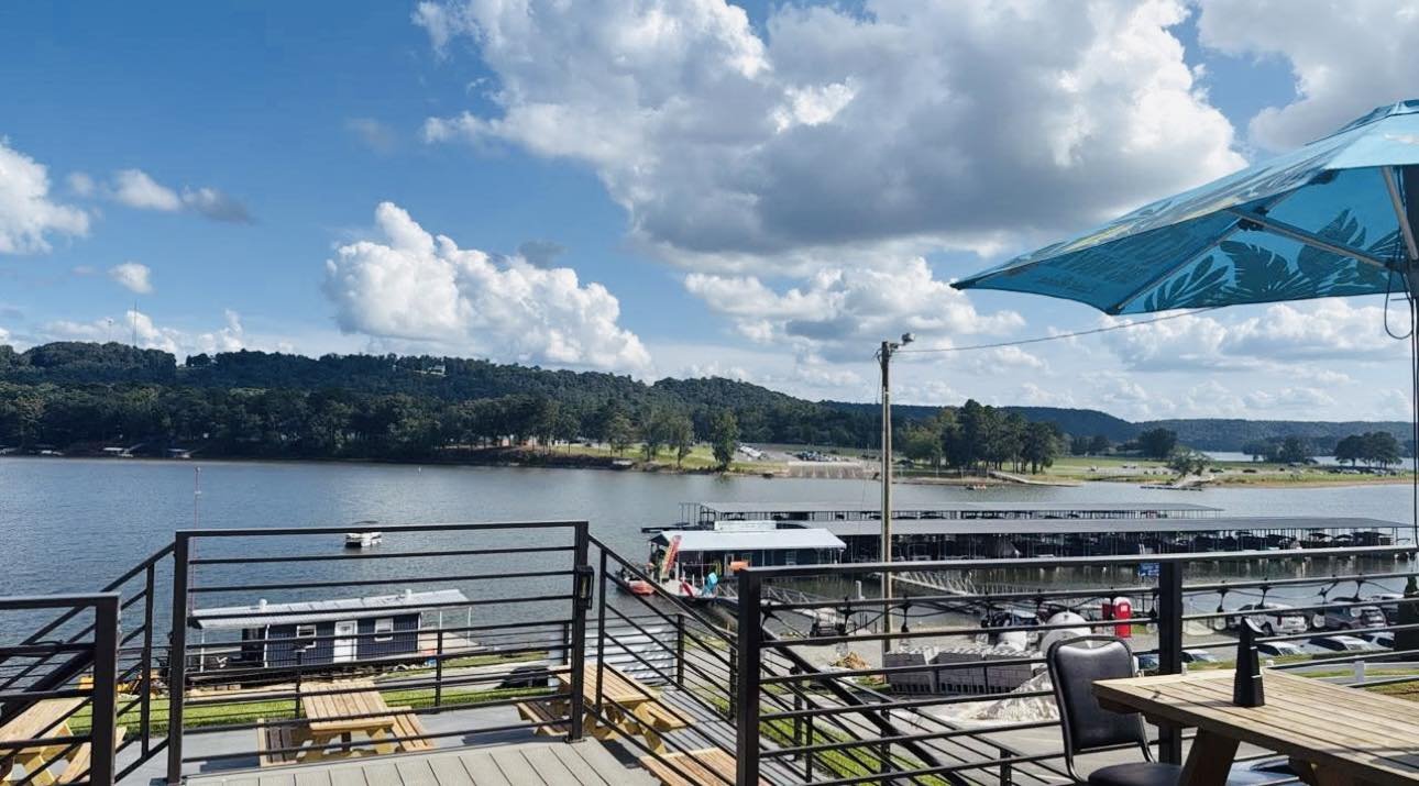 View of a lake with docked boats and a hillside in the background, seen from a balcony with seating and a blue umbrella.