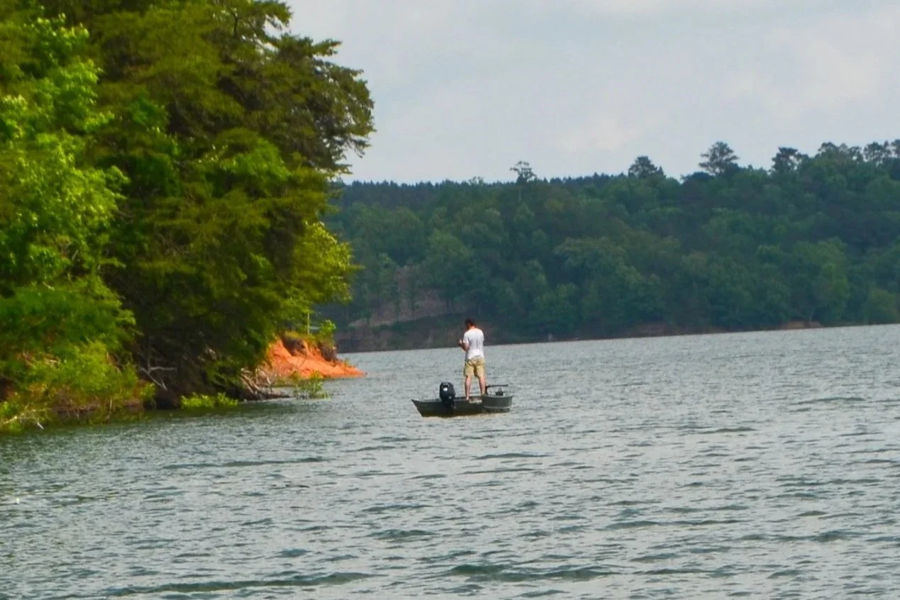 A person standing in a small boat on Smith Lake near the T, fishing near the shoreline with dense trees and wooded landscape in the background.