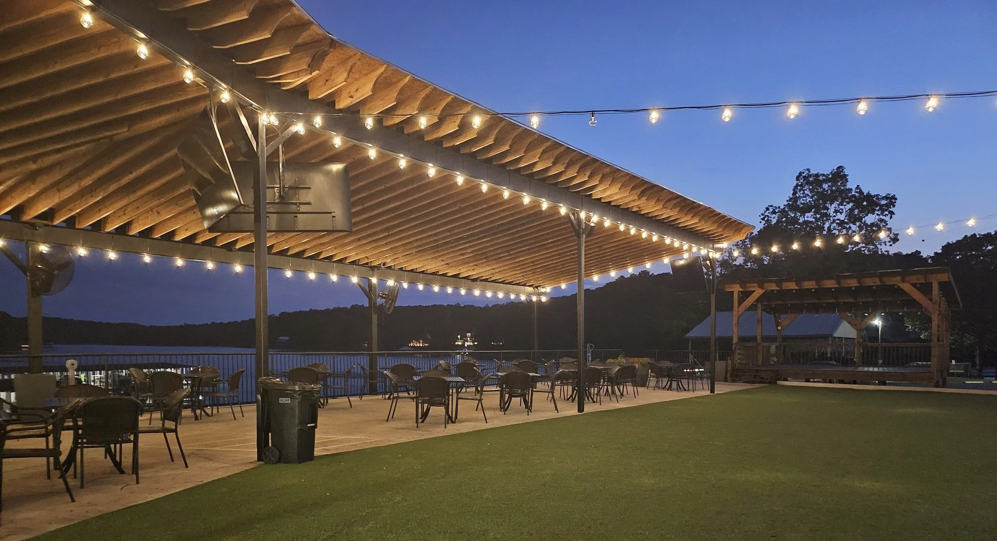 An outdoor patio area with tables and chairs, string lights hanging overhead, overlooking a body of water and trees at twilight.