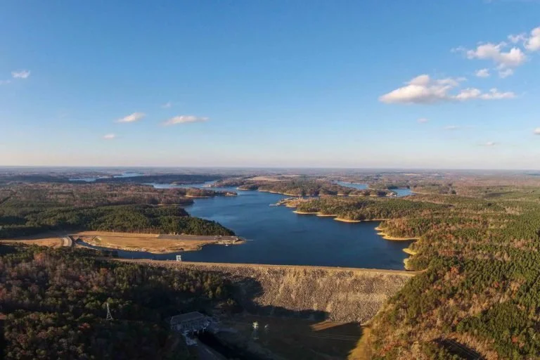 A large reservoir with a dam in the foreground, surrounded by forested hills and a blue sky with some clouds.