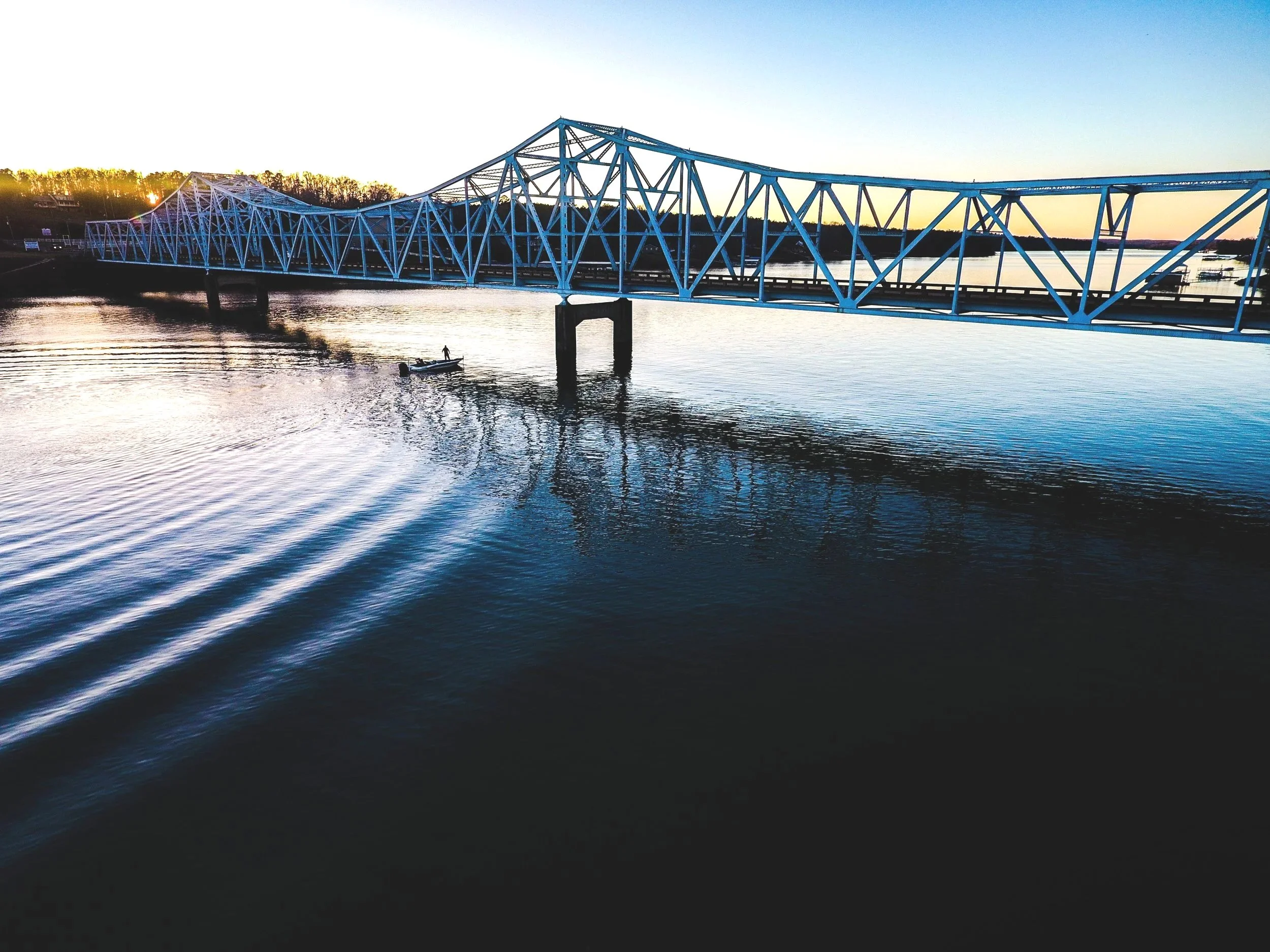 Blue bridge over calm river at sunset with a person in a boat nearby.