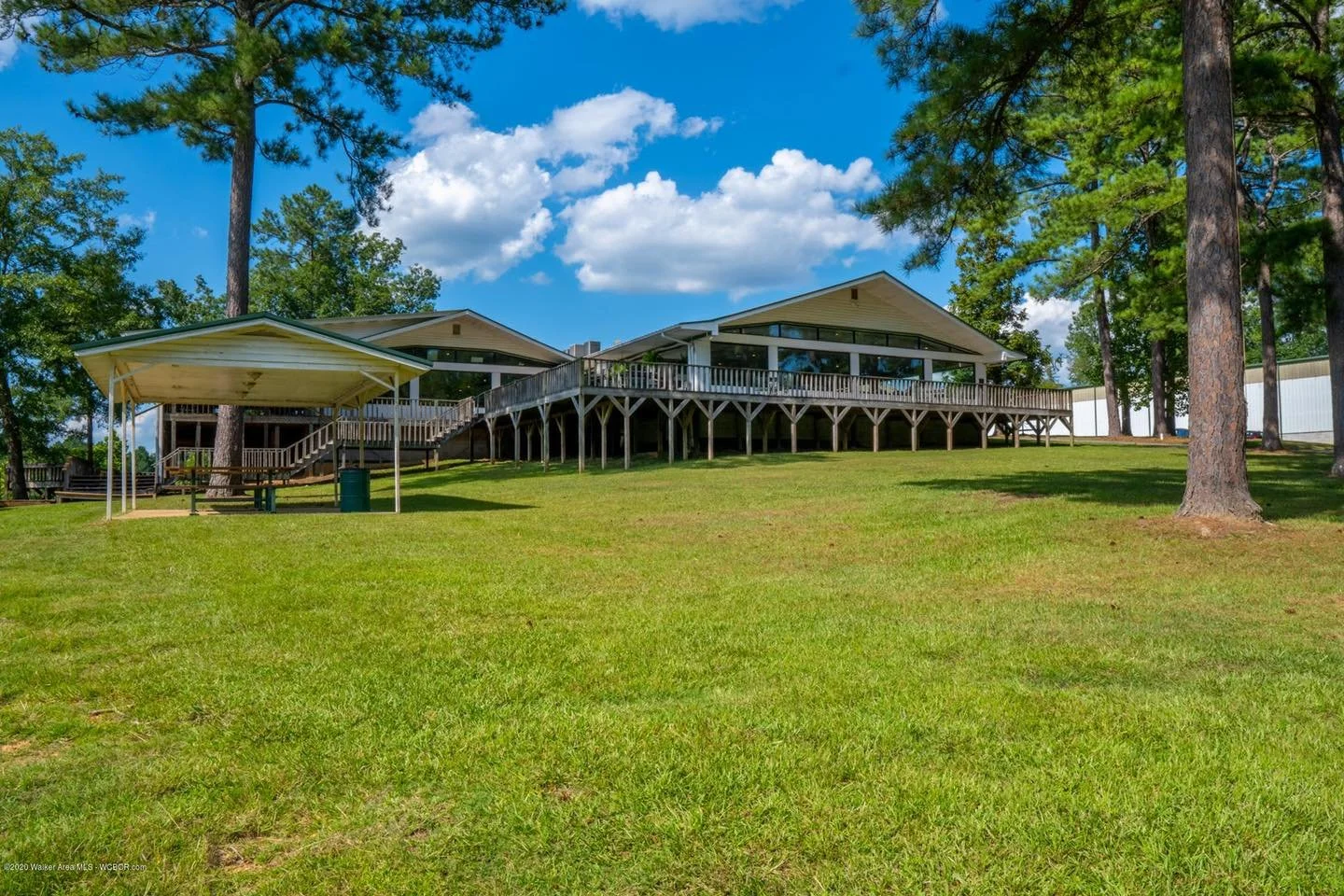 Large house or lodge on a grassy hill with tall pine trees and a bright blue sky with clouds in the background.