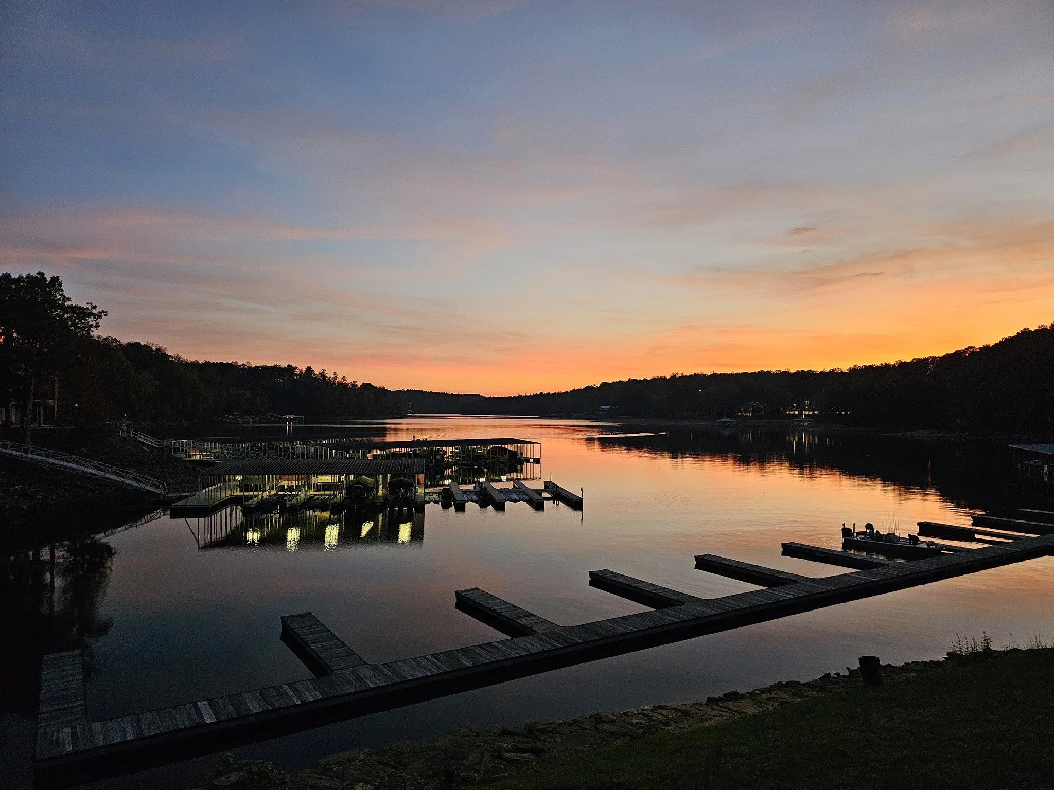 Sunset over a calm lake with docks and boats, silhouetted trees, and a colorful sky with pink, orange, and blue hues.
