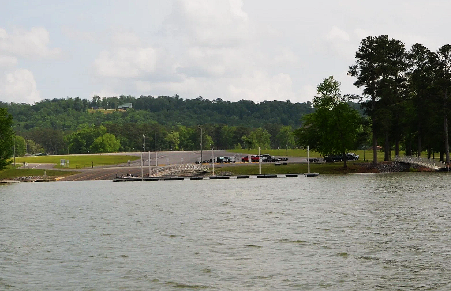 View of Smith Lake with calm water in the foreground, a public boat launch at Smith Lake Park, dock extending into the water, and a parking lot with several vehicles, surrounded by green trees and hills under a partly cloudy sky.