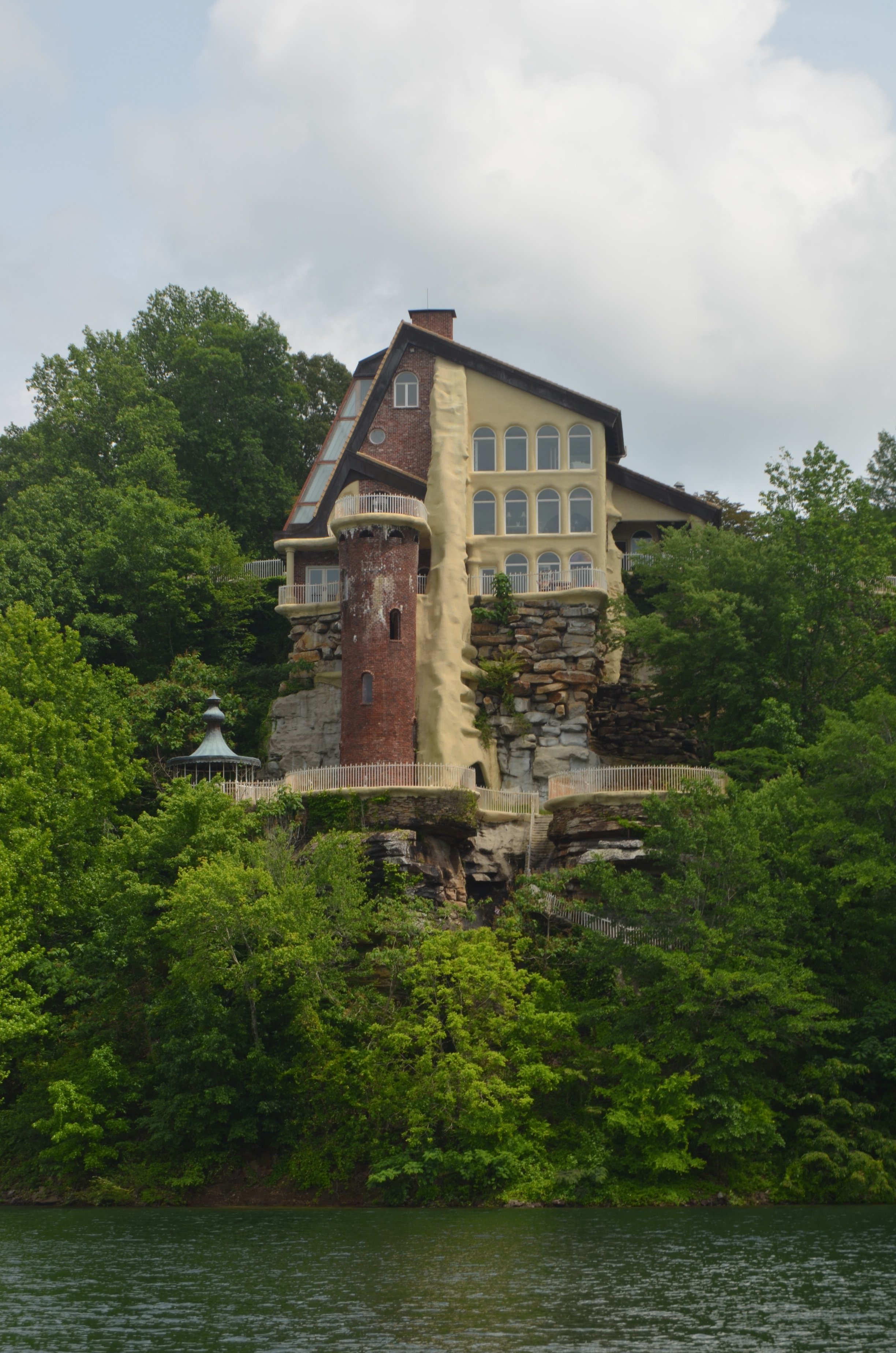 A house built into the side of a hill, featuring multiple rounded windows, surrounded by green trees and overlooking a body of water.