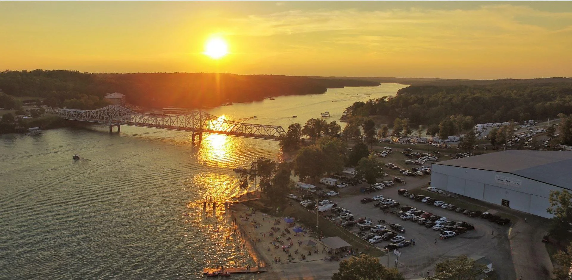 Sunset over a river with boats, a bridge, parked cars, and a beach area with umbrellas and people