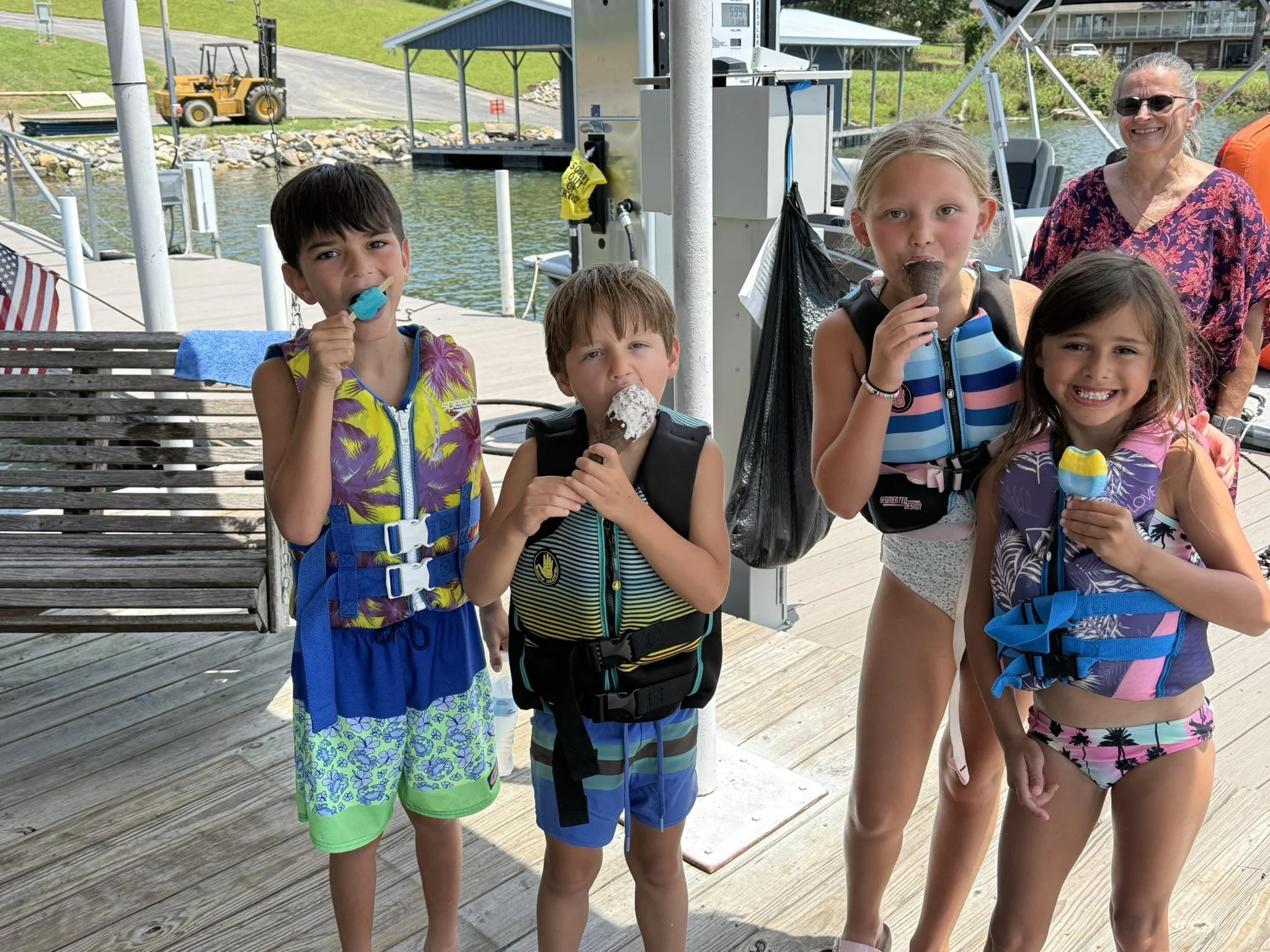 Group of five kids and one adult woman standing on a dock by a boat, eating ice cream and smiling, with a lake, a gazebo, and a yellow construction vehicle in the background.