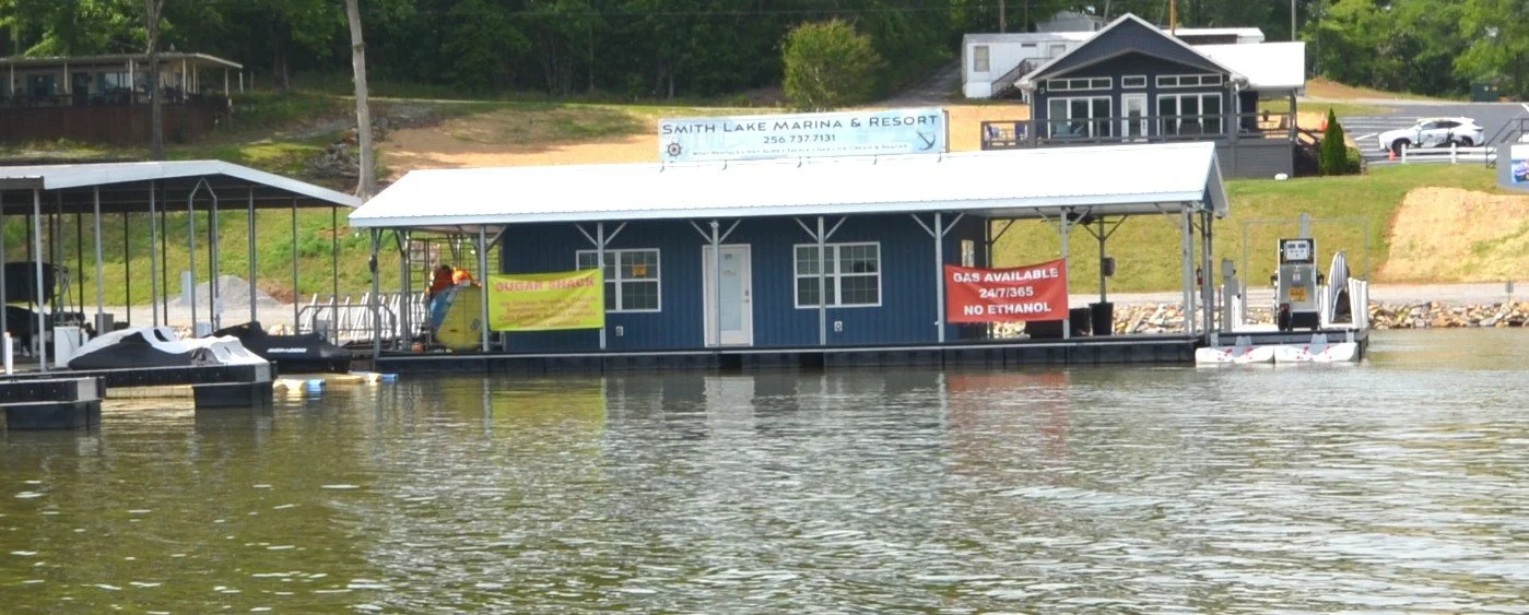 A houseboat docked at a marina with a sign that reads 'Smith Lake Marina & Resort' on the roof, and other signs indicating gas availability and ethanol restrictions. Surrounding water and nearby structures are visible.