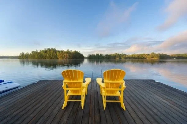 A dock with two yellow Adirondack chairs overlooking a calm lake with trees in the distance under a partly cloudy sky.