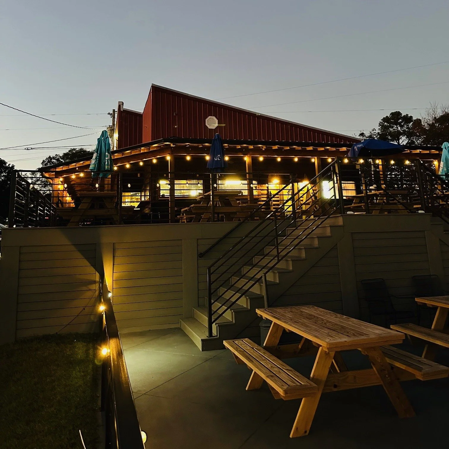 with outdoor seating area illuminated by string lights at dusk. There are picnic tables on the lower level, and a deck with additional seating and umbrellas on the upper level. The building has a red metal roof, and the scene is set against a darkening s
