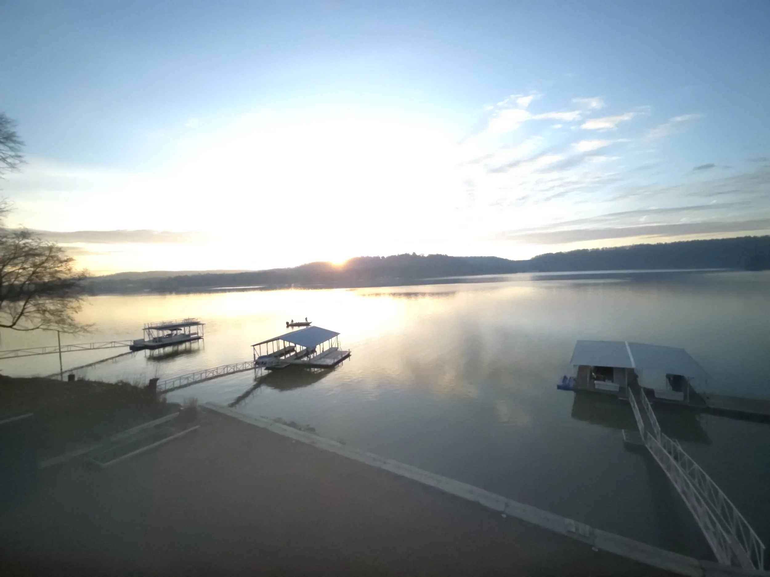Sunrise over a calm lake with a dock and boats, trees on the left side, and a partly cloudy sky.