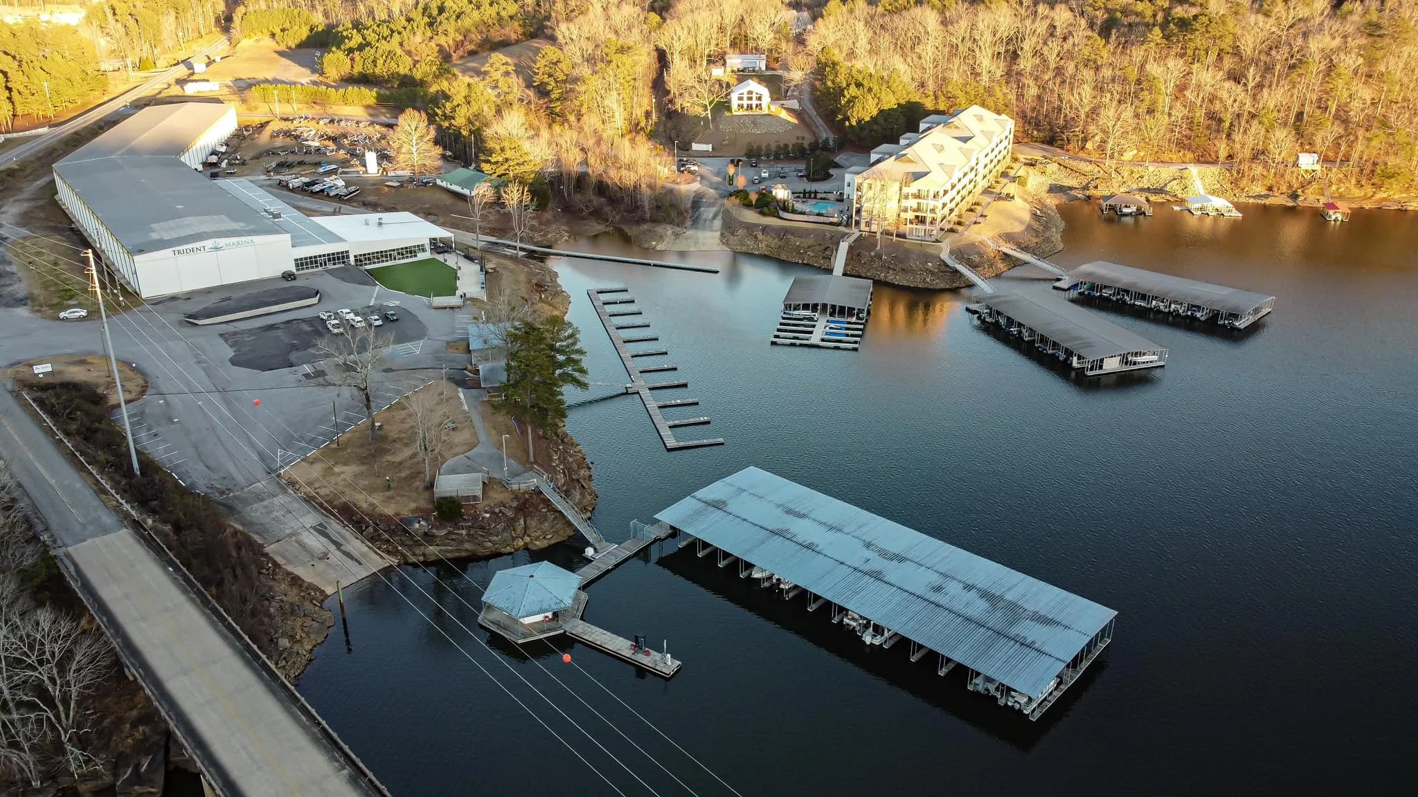 Aerial view of a marina with multiple boat docks on a river, a large building labeled 'Trident Marina', parking lots, and surrounding buildings and wooded area.