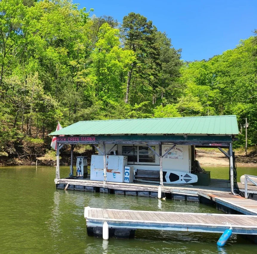 A dockside building at Rock Creek Marina on a sunny day, with a green metal roof, surrounded by lush green trees, and floating on green water.