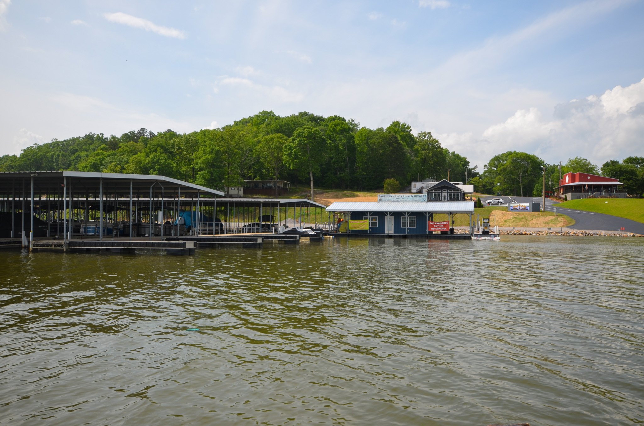 View of a marina with boats docked under covered slips, small buildings along the water, and lush green trees on a hill in the background, with a partly cloudy sky overhead.