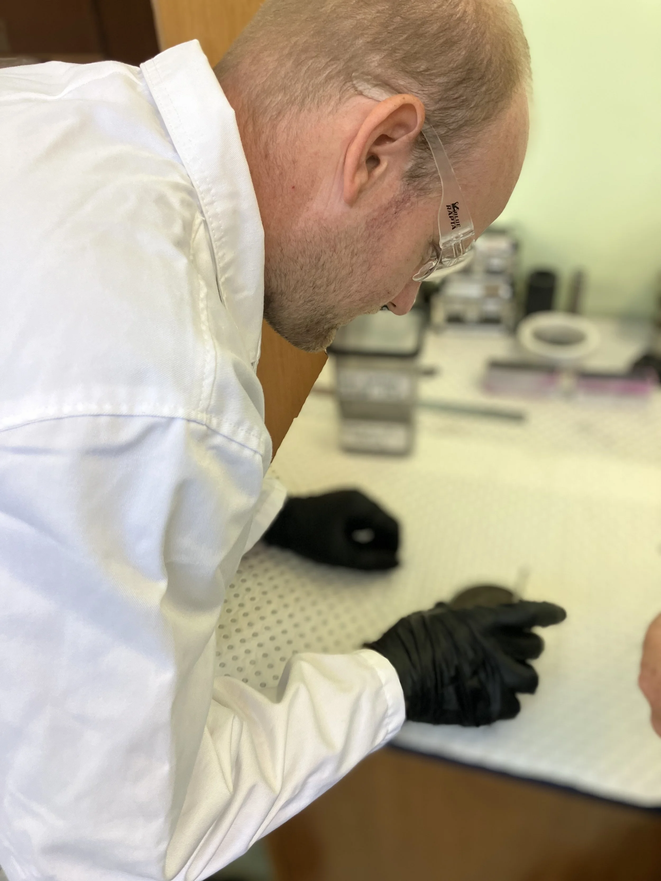 Close-up side view of a man in a white laboratory coat with glasses, seated in a laboratory setting, wearing black gloves, with equipment and a green wall in the background.