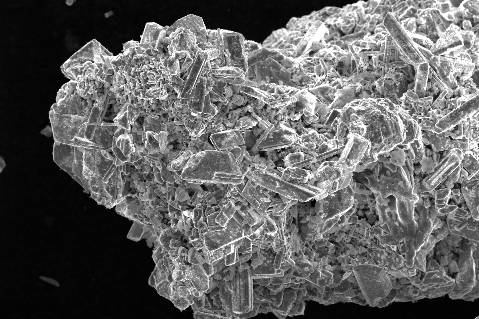 Microscopic view of a crystalline mineral sample, a catalyst, with various angular and elongated crystals against a black background.