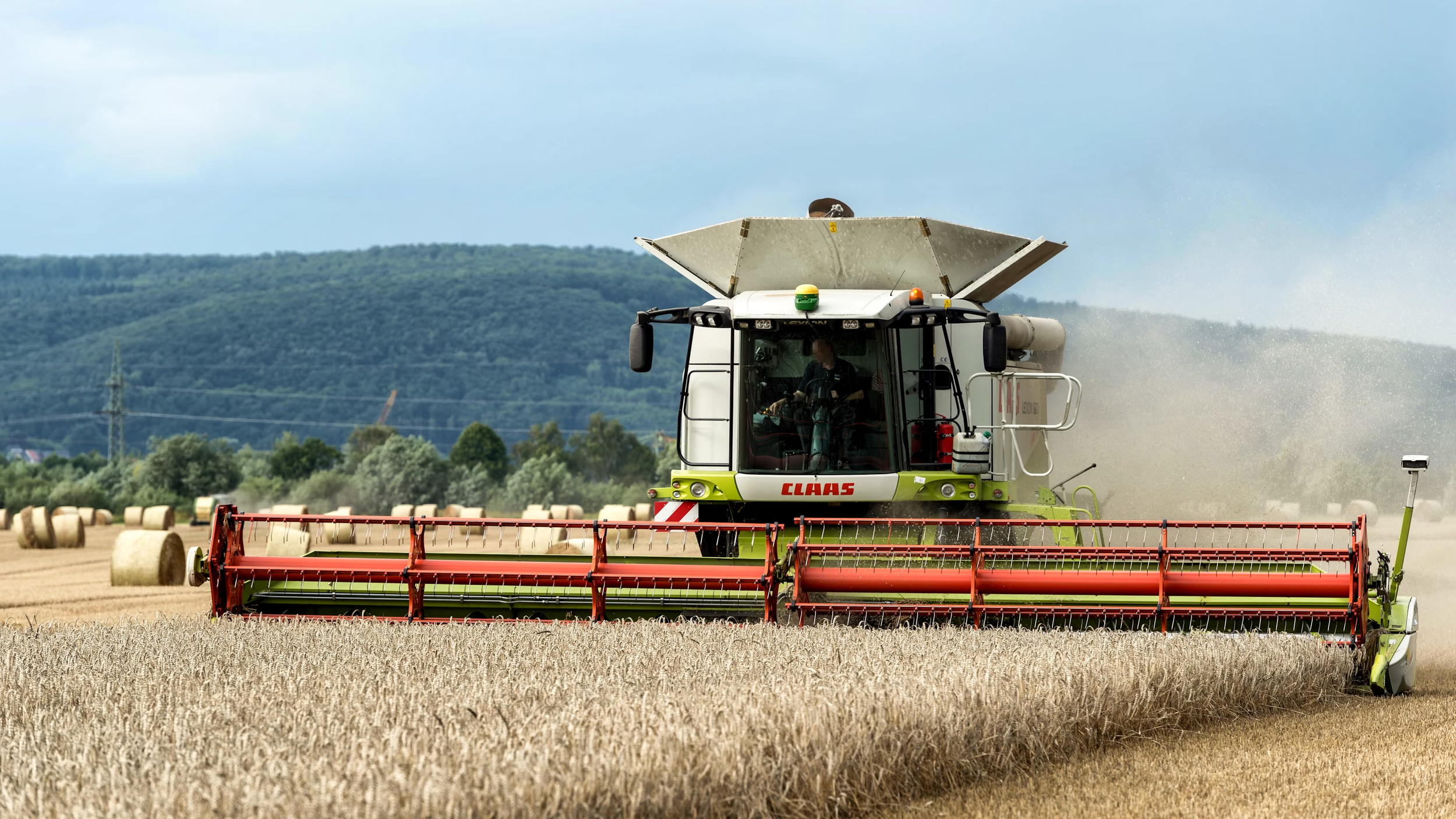 A large CLAAS combine harvester operating in a wheat field during daytime, with hay bales in the background and hills in the distance.