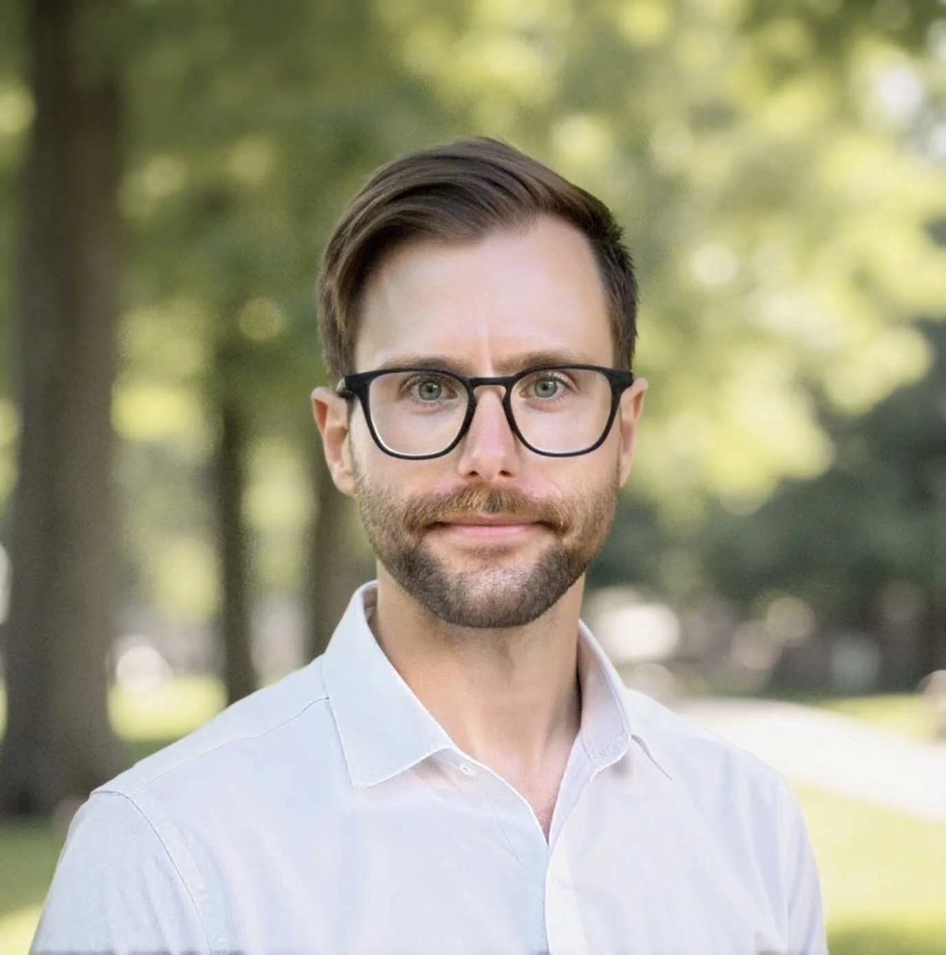 A man wearing glasses and a white shirt outdoors with trees and blurred greenery in the background.