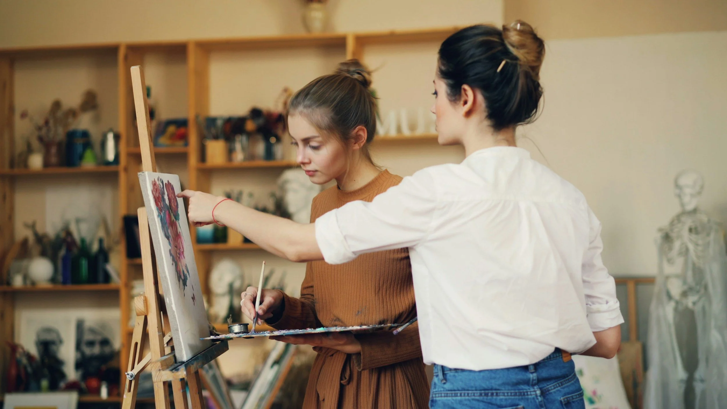 Two young women painting on a canvas in an art studio filled with art supplies and skeleton models in the background.