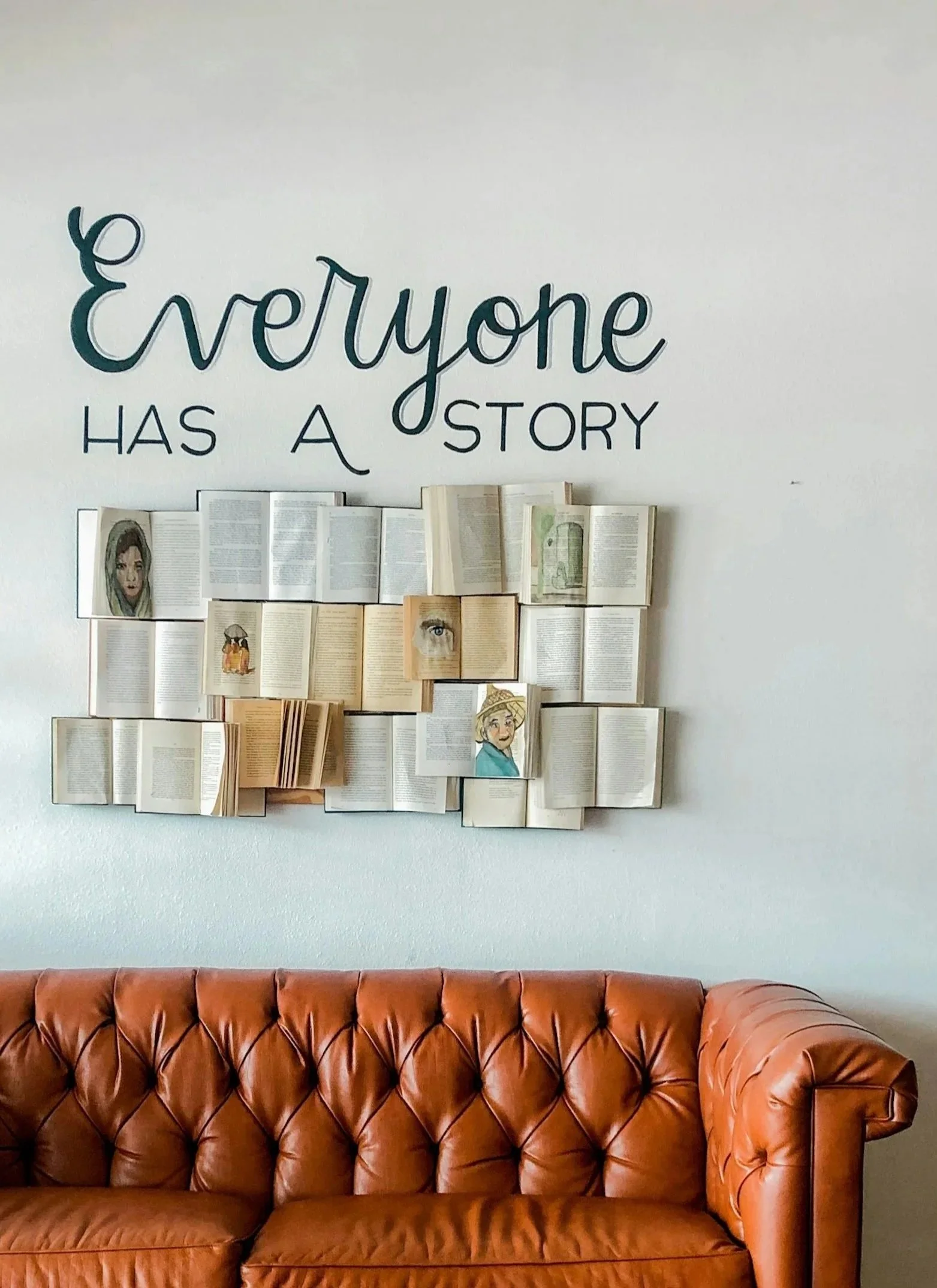 Decorative wall with a quote 'Everyone has a story' and a collage of open books with illustrations, hung above a brown leather tufted sofa.